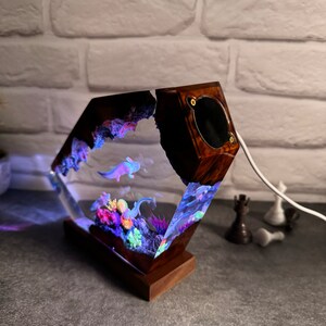 Op de afbeelding: Een houten lampenvoet met een heldere harskubus die een miniatuur onderwater sc&egrave;ne bevat met koraal, planten en axolotls. De harskubus wordt van binnenuit verlicht, waardoor een kleurrijke gloed ontstaat.