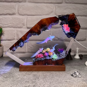 Op de afbeelding: Een harslamp in de vorm van een rotsformatie met een helder, gloeiend interieur. De lamp toont een sc&egrave;ne met axolotls die zwemmen in een koraalrif met groene planten. De lamp wordt van onderen verlicht, waardoor een warm gloed ontstaat.