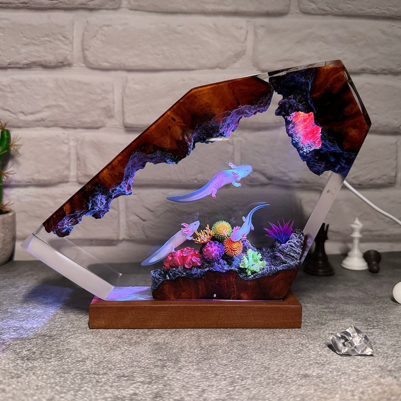 Op de afbeelding: Een harslamp in de vorm van een rotsformatie met een helder, gloeiend interieur. De lamp toont een sc&egrave;ne met axolotls die zwemmen in een koraalrif met groene planten. De lamp wordt van onderen verlicht, waardoor een warm gloed ontstaat.