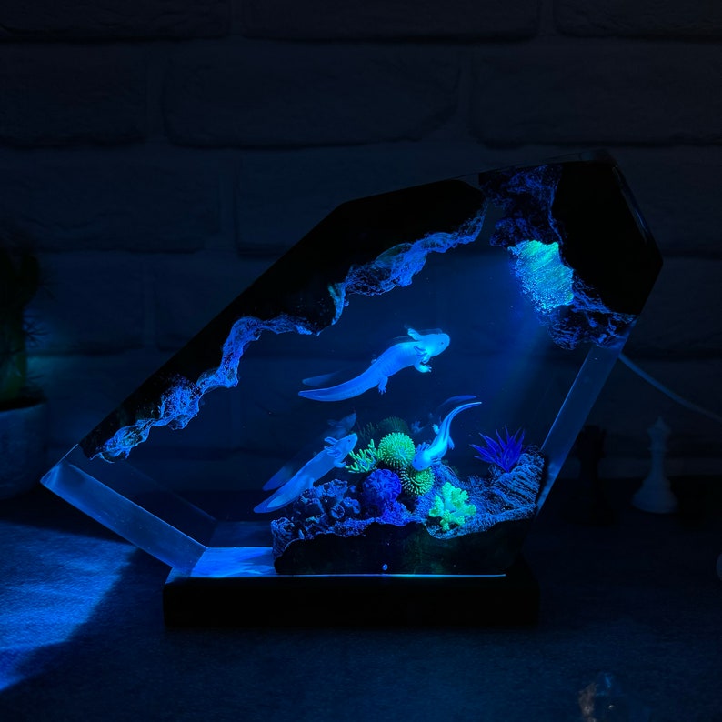 Op de afbeelding: Een transparant harsbeeld van een axolotl, een soort salamander, die zwemt in een blauwe harsbasis. Het beeld wordt van onderen verlicht, waardoor een gloeiend effect ontstaat. De basis is gevormd als een rotsformatie met een grot en planten.