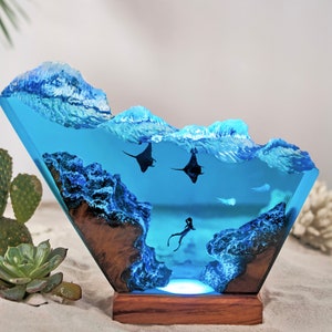 May include: A blue resin sculpture depicting an ocean scene with two manta rays and a mermaid swimming in the water. The sculpture is lit from below, creating a glowing effect.