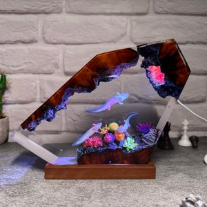 Op de afbeelding: Een harslamp met een houten voet heeft een helder harsblok met een donkerbruin, hout-achtig patroon. Het harsblok bevat drie axolotls en koraalachtige planten. De lamp wordt van onderen verlicht, waardoor de axolotls en planten in een blauwe en paarse gloed worden gezet.