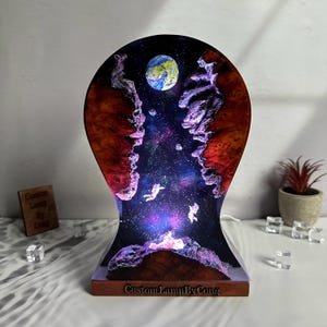 Op de afbeelding: Een houten lampenvoet met een harsinzet die een galaxyscene voorstelt met een blauwe planeet, sterren en astronauten. De voet is donkerbruin van kleur met een lichtbruine houtnerf. De harsinzet is helder en heeft een paarse en blauwe gloed. De lamp is aan en de gloed is zichtbaar. De lampenvoet heeft de tekst "Custom Lamp By Cong" aan de voorkant.