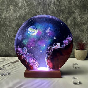 Könnte beinhalten: Eine runde Harzlampe mit einer Galaxie-Szene, mit einem Planeten, Sternen und einem Astronauten. Die Lampe wird von unten beleuchtet und erzeugt ein warmes Leuchten.
