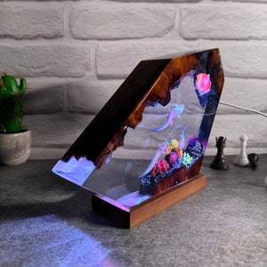 Op de afbeelding: Een harslamp met een houten voet heeft een helder harsgedeelte met een sc&egrave;ne van axolotls die zwemmen in een koraalrif. De lamp wordt van onderen verlicht, waardoor een blauwe gloed ontstaat.
