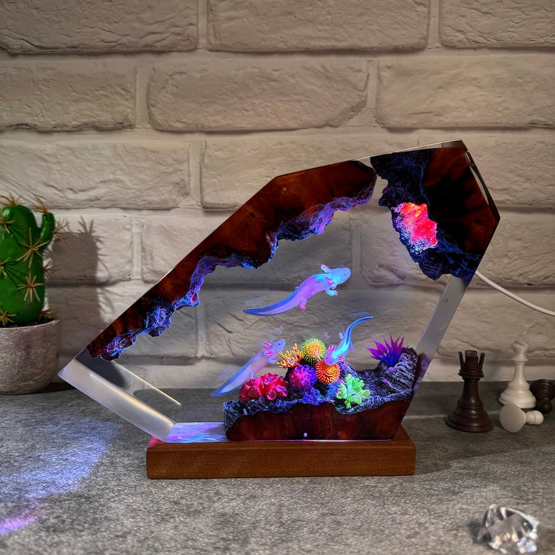 Op de afbeelding: Een harslamp in de vorm van een rotsformatie met een helder interieur. De lamp heeft drie axolotls die in het hars zwemmen, samen met kleurrijke koralen en planten. De basis van de lamp is gemaakt van hout.