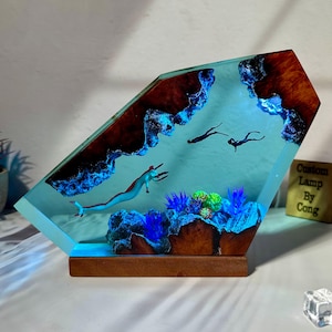 Może przedstawiać: Lampa z żywicy z niebiesko-zieloną sceną podwodną. Lampa przedstawia konika morskiego, koralowce i dwóch nurków. Podstawa lampy jest wykonana z drewna i zawiera napis "Custom Lamp By Cong".