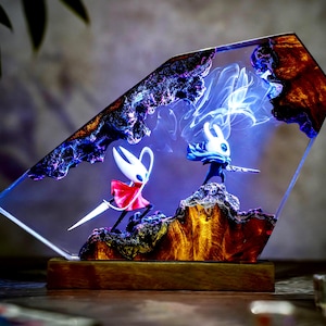 May include: A decorative resin lamp featuring two stylized figures in a dynamic pose, set against a backdrop of swirling blue and white light. The lamp has a wooden base and a clear, angled resin enclosure with a rocky, textured interior.