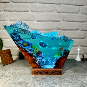 May include: A resin sculpture of an ocean scene with a wave cresting over a coral reef and two sea turtles swimming. The sculpture is lit from below and has a wooden base with the text "CustomLampByCong".