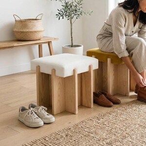 May include: A light-filled room with a woman tying her brown leather boots. Two wooden stools with upholstered tops, one white and one yellow, are in the foreground. Various pairs of shoes are nearby, including sneakers and loafers. A woven rug and a small tree are also visible.