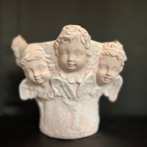 Könnte beinhalten: Ein weißer Keramiktopf in Form von drei Cherubinen mit Flügeln. Der Topf ist hellrosa und hat ein detailliertes Design.
