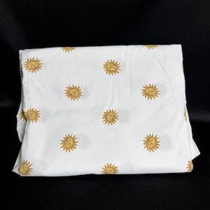 Pode incluir: Um lençol de tecido branco com um padrão repetitivo de sóis amarelo-dourados com traços faciais estilizados. Cada ícone de sol está espaçado uniformemente pela superfície do tecido, criando um design consistente e simétrico em toda a peça.