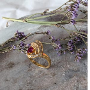 May include: A gold ring featuring a pear-shaped red gemstone at the center, accented by a fan-shaped arrangement of baguette-cut clear stones and a row of small round clear stones along the band, creating a layered and detailed design.
