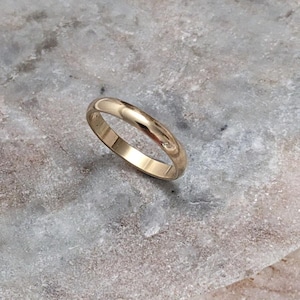 May include: A gold wedding band with a polished finish. The ring is round and sits on a gray, textured surface. The band is a simple, classic design.