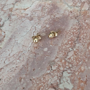 May include: Two small, gold-colored ball stud earrings. The earrings are round and shiny, with a post and backing. The earrings are displayed on a pink and brown marble surface.