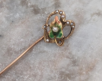 May include: A gold-toned hat pin with a heart-shaped top. The heart is adorned with small pearls and a green and orange floral design. The pin is set against a neutral background.