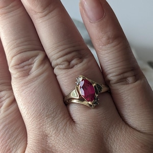 May include: A gold ring featuring a large, oval-cut, deep red gemstone as the centerpiece. The ring has a split shank design with small accent stones on either side of the main stone. The ring is set on a hand.