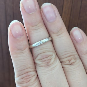 May include: A polished silver-toned band ring featuring a single, small, round, clear gemstone set flush into the center of the metal surface. The ring has a smooth, rounded profile and a simple, clean design that encircles the finger.