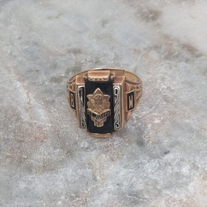 May include: A gold-toned ring with a black center stone. The ring features the text "St. Joseph" and the year "1943". The ring has a rectangular shape with detailed side designs. The ring is set against a light gray, textured surface.