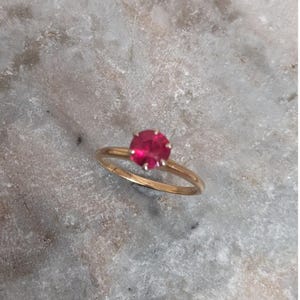 May include: A gold ring featuring a single round, faceted, vibrant pink gemstone set in a prong setting. The band is thin and polished, showcasing a simple design that highlights the central stone's bright color and reflective surface.