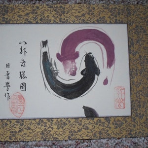 May include: A traditional Japanese painting on rice paper with a black and purple ink design of two fish swimming in a circle. The painting is framed in a gold and black frame with a floral pattern. The text on the painting reads "法隆寺" which translates to "Horyuji Temple".