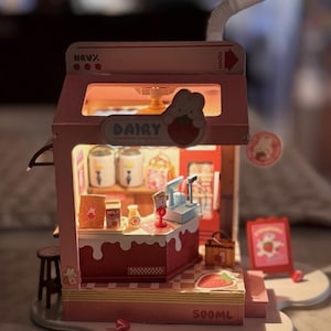May include: A miniature pink and red dairy shop model with a strawberry theme. The model includes a counter, shelves with milk bottles, and a sign that says "DAIRY". A straw extends from the top, and the base reads "500ML".