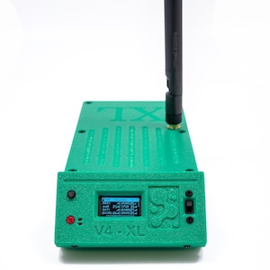 May include: A green electronic device with a black antenna and a small screen displaying data. The device has the text "TX" and "V4-XL" printed on it. It features buttons and a switch on the front.