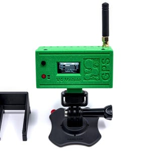 May include: A green GPS device labeled "V4 Mobile" with a small screen, antenna, and a black mounting system. Includes a black bracket and a black base with a red button. The device is designed for mobile use.