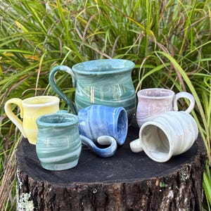 May include: A collection of handmade ceramic mugs in various colors, including yellow, blue, green, and pink. Each mug features a unique marbled design and a curved handle. The mugs are displayed on a tree stump.
