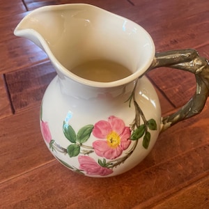 May include: A cream-colored ceramic pitcher featuring a painted design of pink wild roses with green leaves and stems. The pitcher has a wide spout and a textured, dark green handle shaped like a gnarled tree branch, adding a distinct organic element.