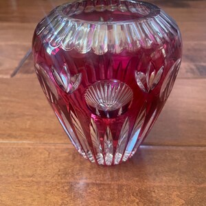 May include: A deep red cut-to-clear crystal vase featuring a wide, circular opening with a scalloped rim. The body of the vase is decorated with intricate geometric patterns, including vertical fluting, circular medallions, and leaf-shaped motifs.