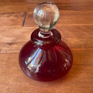 May include: A deep red glass decanter with a wide, rounded base and a narrow neck. The decanter is topped with a clear, spherical glass stopper that sits securely in the opening. The glass has a smooth, polished finish with reflective surfaces.