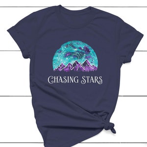 以下が含まれることがあります： 紺色のTシャツに、水彩画で描かれた山と星空のデザインが施されています。デザインの下には、「CHASING STARS」という文字が白で印刷されています。