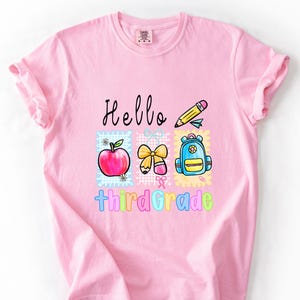 Pode incluir: Camiseta rosa claro com as palavras "Hello third grade" em letras pretas e arco-íris. A camiseta apresenta ilustrações coloridas de uma maçã, um lápis, um laço e uma mochila.