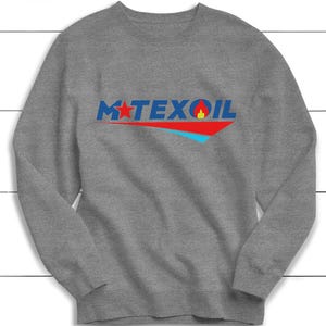 以下が含まれることがあります： グレーのスウェットシャツ。青色の「M TEXOIL」の文字、赤い星と炎のグラフィックが特徴です。テキストの下には赤と青のデザインがあります。クルーネックのスタイルです。