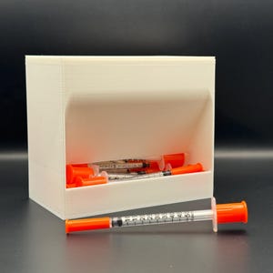 May include: A white, rectangular container with an open front, holding several syringes with orange caps. One syringe rests in front of the container. The syringes have clear barrels with black measurement markings.