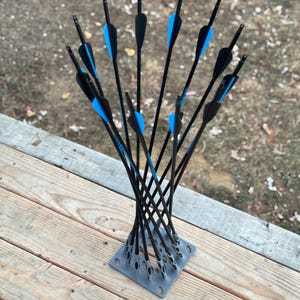 May include: A collection of black arrows with blue fletching, arranged in a gray square base. The arrows are standing upright, creating a visually appealing display. The background is a wooden deck and a blurred outdoor setting.