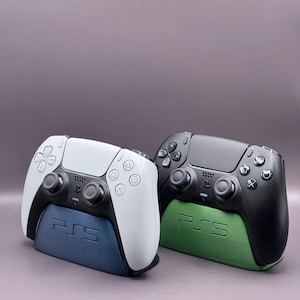 May include: Two PlayStation 5 controllers, one white and one black, are displayed on matching charging stands. The stands are labeled "PS5" and are colored blue and green.