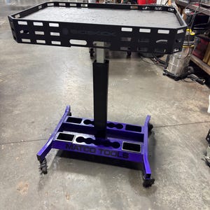 May include: A black and purple rolling tool cart with the text "SIDEKICK" and "MATCO TOOLS". The cart has a rectangular top with cut-out details and a purple base with tool holder slots. It is designed for workshop use.