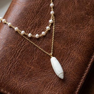 Puede incluir: Un collar de varias capas que presenta una cadena dorada con pequeñas perlas blancas y una segunda cadena dorada con un colgante de concha blanca alargada. El colgante tiene una forma lisa y cónica con sutiles estrías en la base.