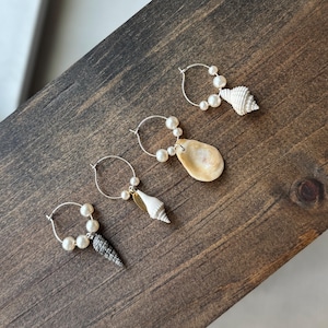 May include: Four silver hoop earrings, each adorned with small white pearls. Three of the hoops feature a single seashell charm, including a white spiral shell, a flat tan shell, and a textured grey cone-shaped shell hanging from the metal wire.