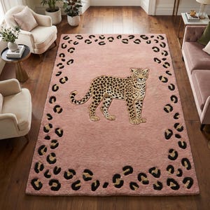 Peut inclure: Un tapis rectangulaire au fond rose tendre orné d'une illustration centrale représentant un léopard debout. Le tapis est décoré d'une bordure composée de taches de léopard noires et marron foncé, formant un cadre autour de l'animal.