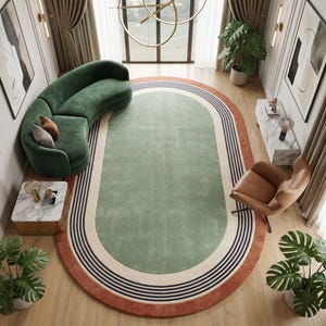 Könnte beinhalten: Ein ovaler Teppich mit einem zentralen salbeigrünen Feld, umgeben von konzentrischen Streifen in Creme, Dunkelblau und einem terrakottafarbenen Rand. Der Teppich liegt auf einem hellen Holzboden in einem Raum mit geschwungenem grünem Sofa.