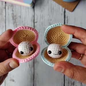 May include: Two crocheted clam shells with smiling pearl faces. One shell is pink and the other is blue.