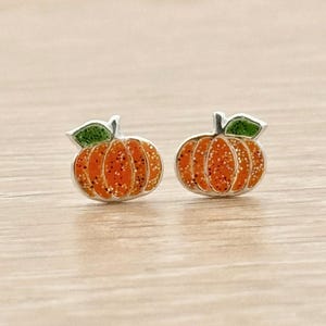 Könnte beinhalten: Ein Paar Ohrstecker in Kürbisform. Jeder Ohrstecker hat einen leuchtend orangefarbenen Körper mit glitzerndem Finish und ein kleines grünes Blatt an der Oberseite. Das Kürbisdesign ist mit einem metallisch silbernen Rand versehen.