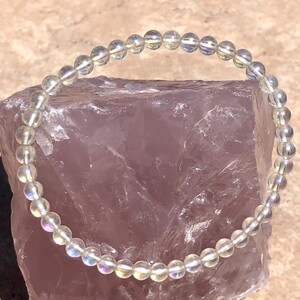 May include: A white iridescent gemstone bracelet with round beads. The bracelet is on a pink background.