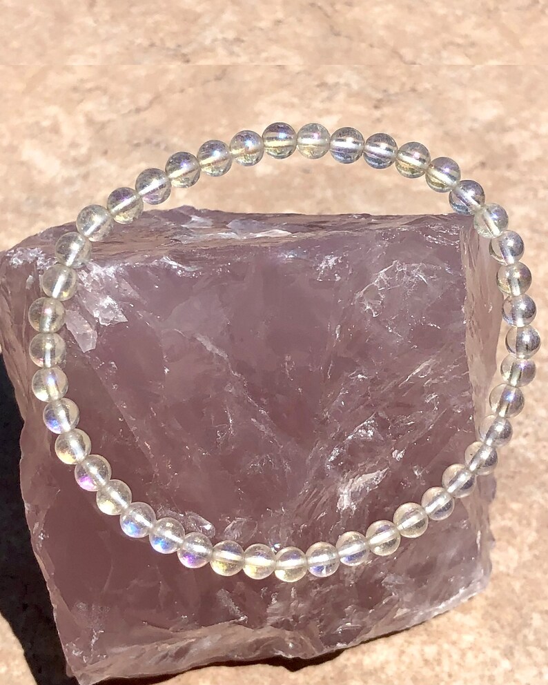 May include: A white iridescent gemstone bracelet with round beads. The bracelet is on a pink background.