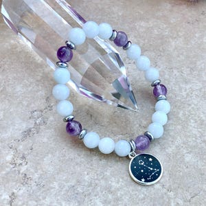 Könnte beinhalten: Ein weißes und violettes Perlenarmband mit einem silbernen Anhänger, der die Konstellation Zwillinge zeigt. Das Armband besteht aus weißen Mondsteinperlen, Amethystperlen und silbernen Zwischenstücken.