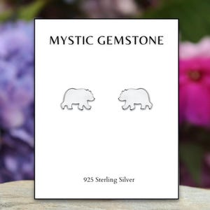 May include: White bear-shaped stud earrings on a white card with the text "MYSTIC GEMSTONE" and "925 Sterling Silver". The earrings are set against a blurred background of purple and pink flowers.