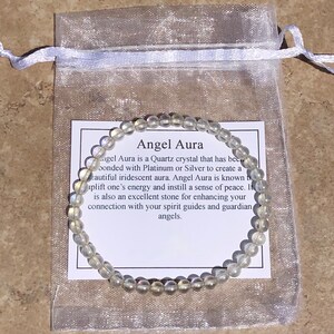 May include: A white Angel Aura bracelet made of clear quartz crystals with iridescent rainbow hues. The bracelet is displayed on a white organza bag with a card that reads "Angel Aura is a Quartz crystal that has been bonded with Platinum or Silver to create a beautiful iridescent aura. Angel Aura is known to uplift one's energy and instill a sense of peace. It is also an excellent stone for enhancing your connection with your spirit guides and guardian angels."